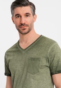 Olivově zelené tričko s výstřihem do V vyrobené z měkké tkaniny, s náprsní kapsou a kontrastními tmavě zelenými ohrnutými rukávy. Hladká textura, volný střih.