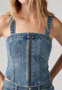 Kvinna som bär en ärmlös blå denimkorsett med dragkedja i fram, tillsammans med matchande denimjeans.