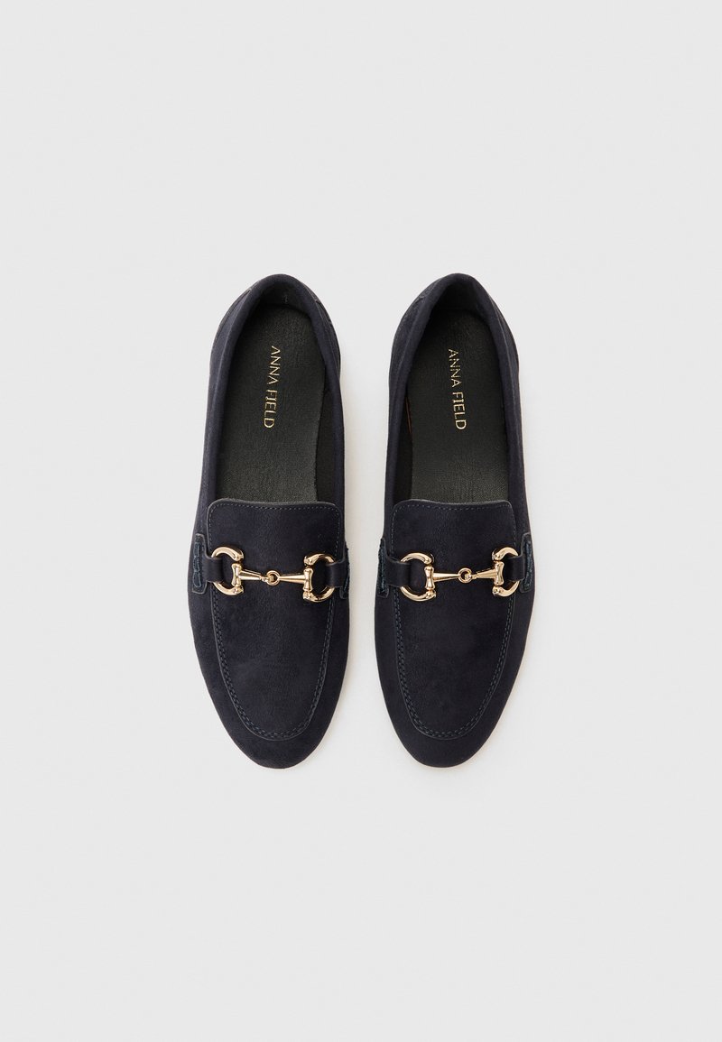 Mocasines de ante azul marino con detalles de hardware en tono dorado, punta redondeada y costuras a lo largo de los bordes. Textura suave y diseño fácil de poner.