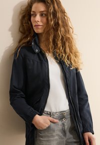 Marineblauw lichtgewicht jack met ritssluiting, drukknoopkraag, zijzakken en elastische manchetten, gecombineerd met een witte top en grijze broek.