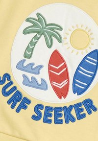 Sárga textil, amely hímzett mintázattal rendelkezik: egy pálmafa, nap, két szörfdeszka (piros és kék) és hullámok. Szöveg: "SZÖRF KERESŐ."