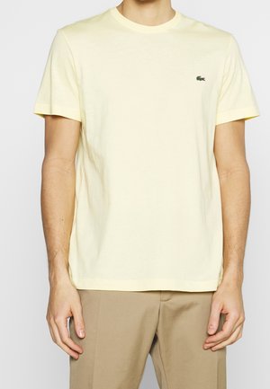 Homme portant un t-shirt jaune clair uni à manches courtes avec un petit logo crocodile vert sur la poitrine, associé à un pantalon beige.