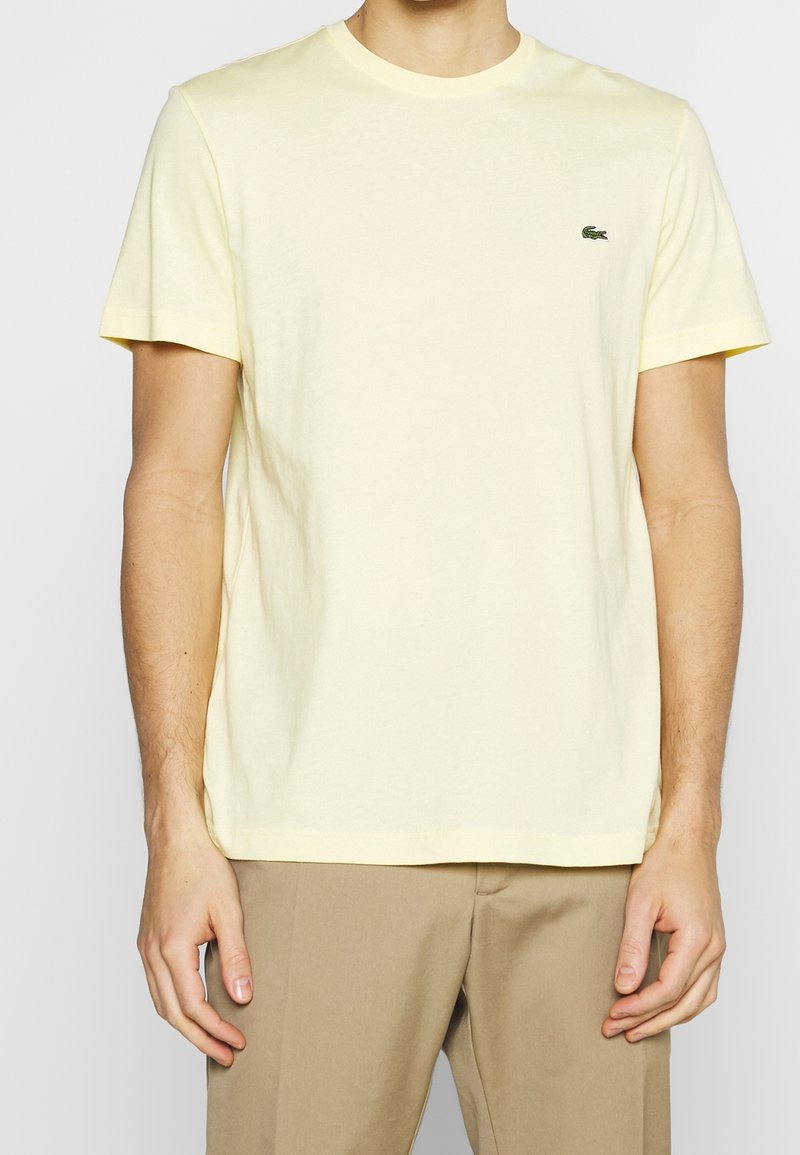 Homme portant un t-shirt jaune clair uni à manches courtes avec un petit logo crocodile vert sur la poitrine, associé à un pantalon beige.