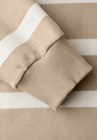 Beige-weiß gestreifter Stoff mit einem gerippten, beigen Bündchen, das umgeschlagen ist und Strickstruktur sowie Nahtdetails zeigt.