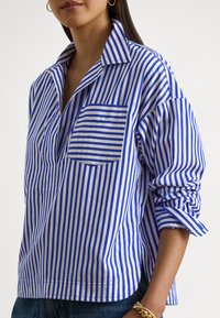 Blau-weiß gestreiftes Hemd mit großem Kragen, Brusttasche und hochgekrempelten Ärmeln; hergestellt aus einem glatten Stoff mit lockerer Passform.