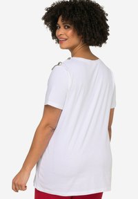 Hvid t-shirt med korte ærmer og rund halsudskæring, med dekorative sølvdetaljer på skuldrene. Glat bomuldsstruktur.