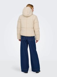 Casaco puffer bege com capuz, apresentando um design matelassé e um corte amplo, combinado com jeans de ganga azul de pernas largas.