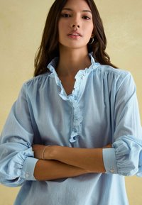 Svetlomodrá pruhovaná blúzka s volánovým golierom a manžetami, dlhé rukávy, gombíkový detail na zápästiach a mäkká, ľahká látka.