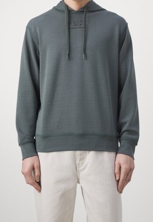 Homme portant un sweat à capuche vert foncé avec cordons et logo discret sur la poitrine, associé à un pantalon beige clair, debout devant un fond uni.