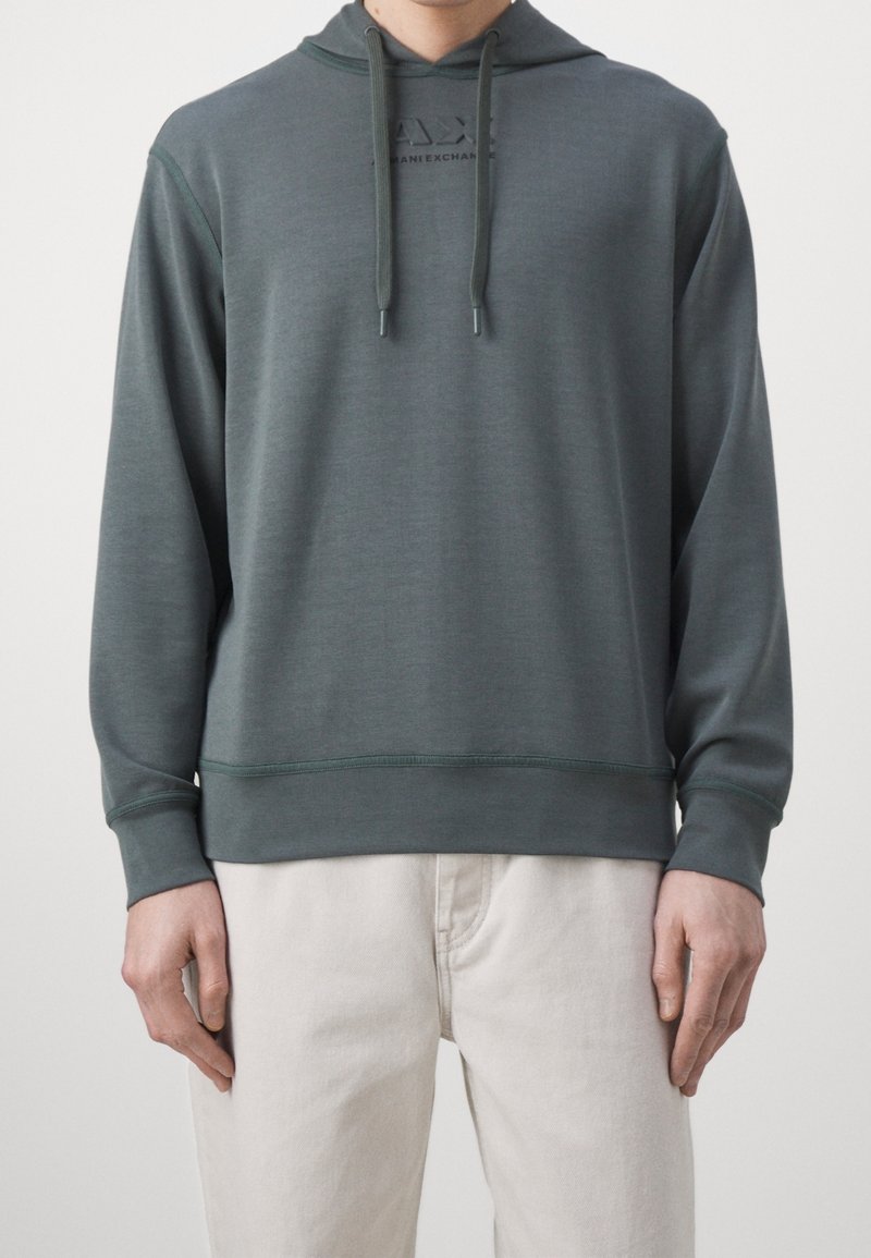 Homme portant un sweat à capuche vert foncé avec cordons et logo discret sur la poitrine, associé à un pantalon beige clair, debout devant un fond uni.