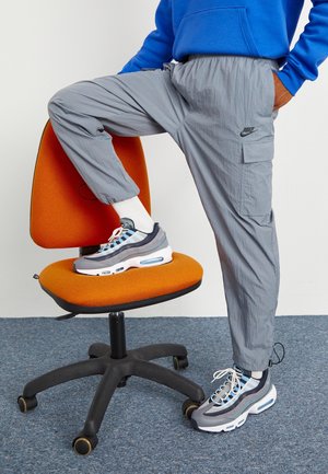 Osoba ubrana w szare spodnie Nike typu cargo oraz szaro-niebieskie buty sportowe z białymi skarpetkami, stojąca jedną stopą na pomarańczowym krześle biurowym.