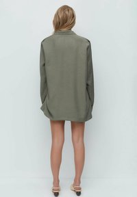 Woman with long blonde hair wearing an oversized olive green blazer, beige heeled sandals, standing facing a light gray wall.