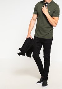 Man i olivgrön polo och svarta slim jeans, håller en svart jacka i ena handen och solglasögon nära kragen med den andra.