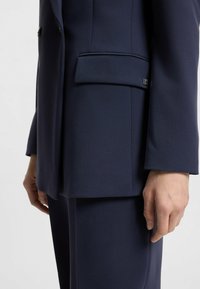 Tummansininen muotolitteinen blazer, joka on valmistettu sileästä kankaasta. Vaate sisältää läpälaput, yhden rintataskun ja hienovaraisia ompeluyksityiskohtia.