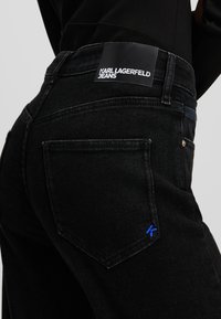 Jeans de denim negro con un parche de logo visible, acentos de costura azul y diseño de bolsillo tradicional. Tejido texturizado con un ajuste ceñido.