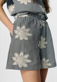 Svart och vit rutig shorts med intrikata beige blommönster, elastisk midja och sidofickor. Mjuk tygstruktur.