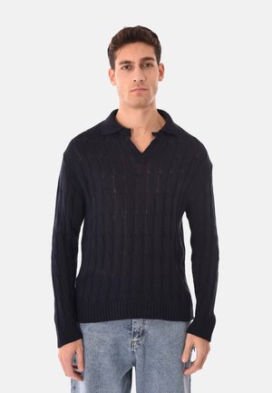 Uomo che indossa un maglione blu navy a trecce con colletto e jeans azzurri, in piedi davanti a uno sfondo bianco semplice.