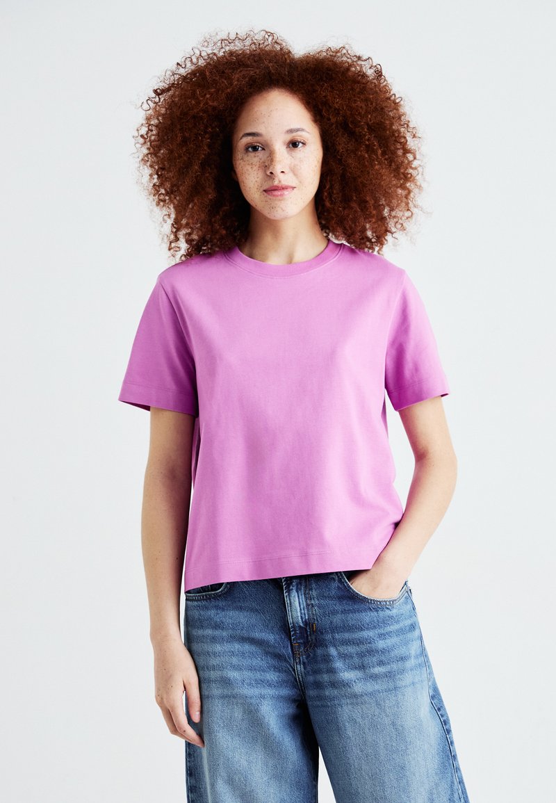 T-shirt in cotone rosa con maniche corte, collo rotondo, vestibilità rilassata e colore solido. Abbinato a jeans in denim blu chiaro, aderenti e svasati.