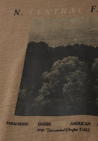 Tryckt tyg som visar ett stiliserat skogslandskap med texten "FARM FRESH", "GOODS" och "AMERICAN CALI" på en brun bakgrund.