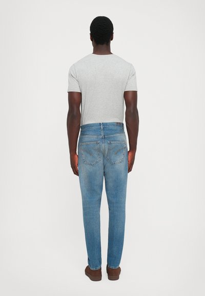 Uomo in piedi di spalle, che indossa una maglietta grigia chiara, jeans blu e scarpe marroni, su uno sfondo chiaro e semplice.