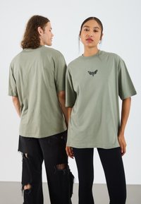 Olive zelené bavlnené oversized tričká s grafickým motýľom v čiernej farbe, vystavené na dvoch jednotlivcoch. Jeden má na sebe čierne roztrhnuté džínsy, druhý čierne legíny.