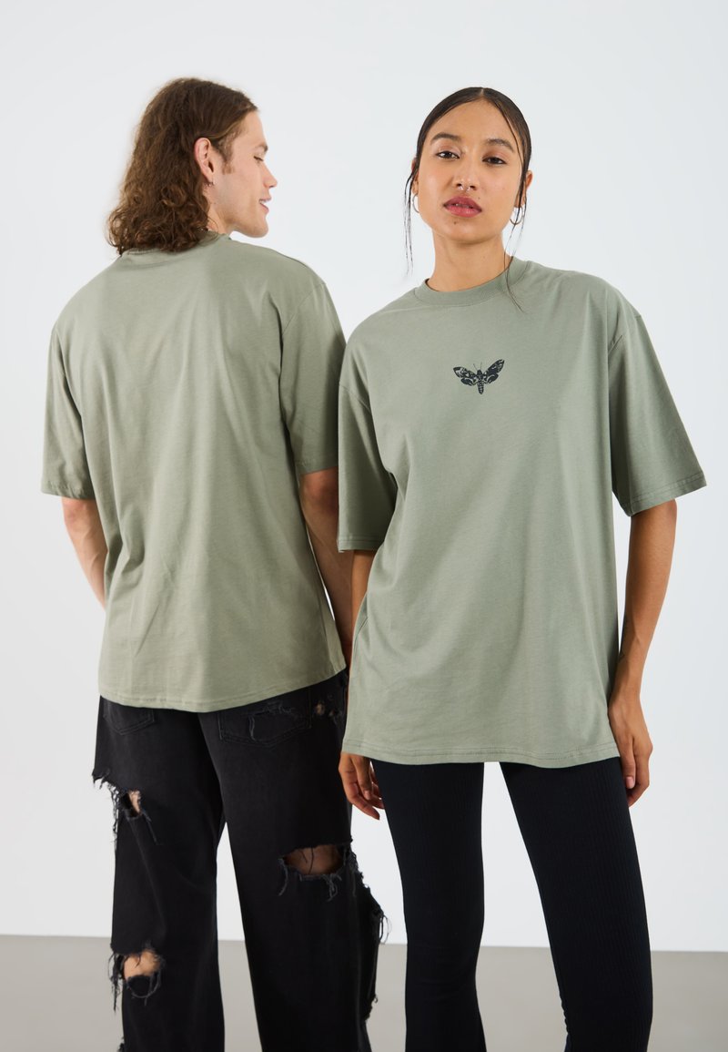 Olive zelené bavlnené oversized tričká s grafickým motýľom v čiernej farbe, vystavené na dvoch jednotlivcoch. Jeden má na sebe čierne roztrhnuté džínsy, druhý čierne legíny.