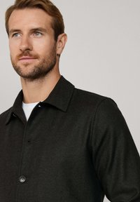 Donkergroen vissengraatjacket met een gestructureerde kraag, een knoopsluiting en een textuurstof, gedragen over een wit T-shirt.