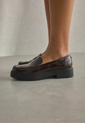 Glänzende dunkelbraune Lederloafer mit dicken schwarzen Gummisohlen, getragen an nackten Füßen, stehend auf glattem grauem Boden.