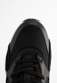 Schwarze Sneaker mit einem strukturierten Mesh-Obermaterial, glatten Lederakzenten und einer weißen Gummisohle. Schwarze Schnürsenkel und ein blauer Akzent an der Spitze.