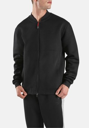 Man draagt een zwarte Reebok-jas met ritssluiting en een zwarte broek met een grijze zijstreep, staat met zijn handen nabij zijn zakken en kijkt naar rechts.