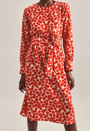 Robe à pois rouges et blancs, manches longues, encolure ronde, taille ceinturée, longueur genou, texture de tissu douce, avec des accents froncés à la taille.