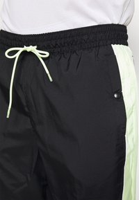 Schwarze Sportshorts mit elastischem Bund und hellgrünem Kordelzug, ausgestattet mit hellgrünen Seitstreifen und Details mit Druckknopf-Taschen.