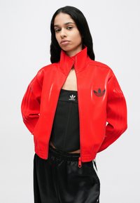 Rdeča bleščeča bomber jakna z visokim ovratnikom, stranskimi zadrgami in črnim logotipom Adidas na prsih. Nošena čez črno majico brez rokavov.