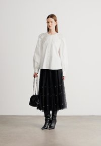 Biela blúzka s ozdobným golierom, čierna tylová sukňa s perličkovými ozdobami a čierne členkové čižmy na podpätku. Modelka drží čiernu tašku.