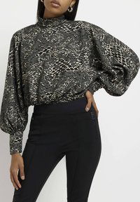 Blouse noire à motif serpent avec un col haut et des manches bouffantes voluminaires, assortie à un pantalon noir ajusté avec un détail de zip sur le côté.