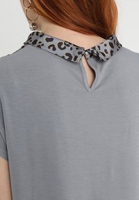Top gris con cuello de estampado de leopardo, mangas cortas y detalle de abertura en la espalda. La tela parece suave con una textura lisa.