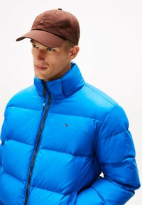 Helderblauwe pufferjack met een hoge kraag, voorzien van horizontaal gestikte secties en een rits aan de voorkant. Bruine pet met logo-details.