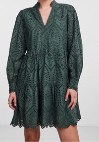 Grünes, knieumspielendes Kleid aus leichtem Material mit filigranen Ausschnittmustern, V-Ausschnitt, langen Ärmeln und gewelltem Saum.