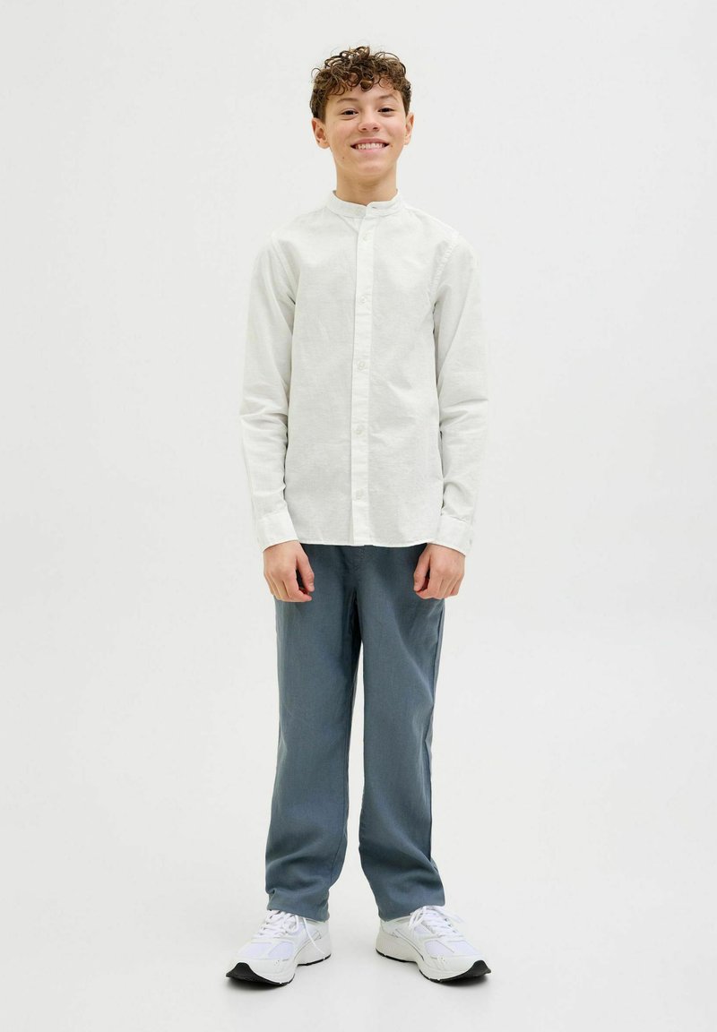 Camicia chiara con colletto alla mandarina, abbinata a pantaloni blu e scarpe da ginnastica bianche, su uno sfondo semplice.