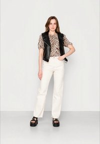 Colete de couro preto sobre uma blusa com estampado de zebra castanho e branco, combinado com calças brancas de perna reta e sandálias de plataforma pretas.