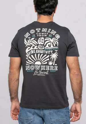 Homme de dos portant un T-shirt noir avec un motif blanc et turquoise de plantes tropicales, du soleil et le texte : « Rien ne vient de nulle part. Définissez la créativité. »