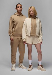 Felpa con cappuccio color talpa e pantaloni su un uomo, bomber beige e pantaloni corti su una donna. Entrambi indossano sneaker neutre. Tessuti morbidi, dettagli minimi.