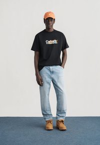 Μαύρος άνδρας όρθιος, φοράει πορτοκαλί καπέλο, μαύρο μπλουζάκι με σχέδιο, ανοιχτό μπλε τζιν και ταμπά εργαστηριακά μποτάκια σε μπλε χαλιά με ουδέτερο φόντο.