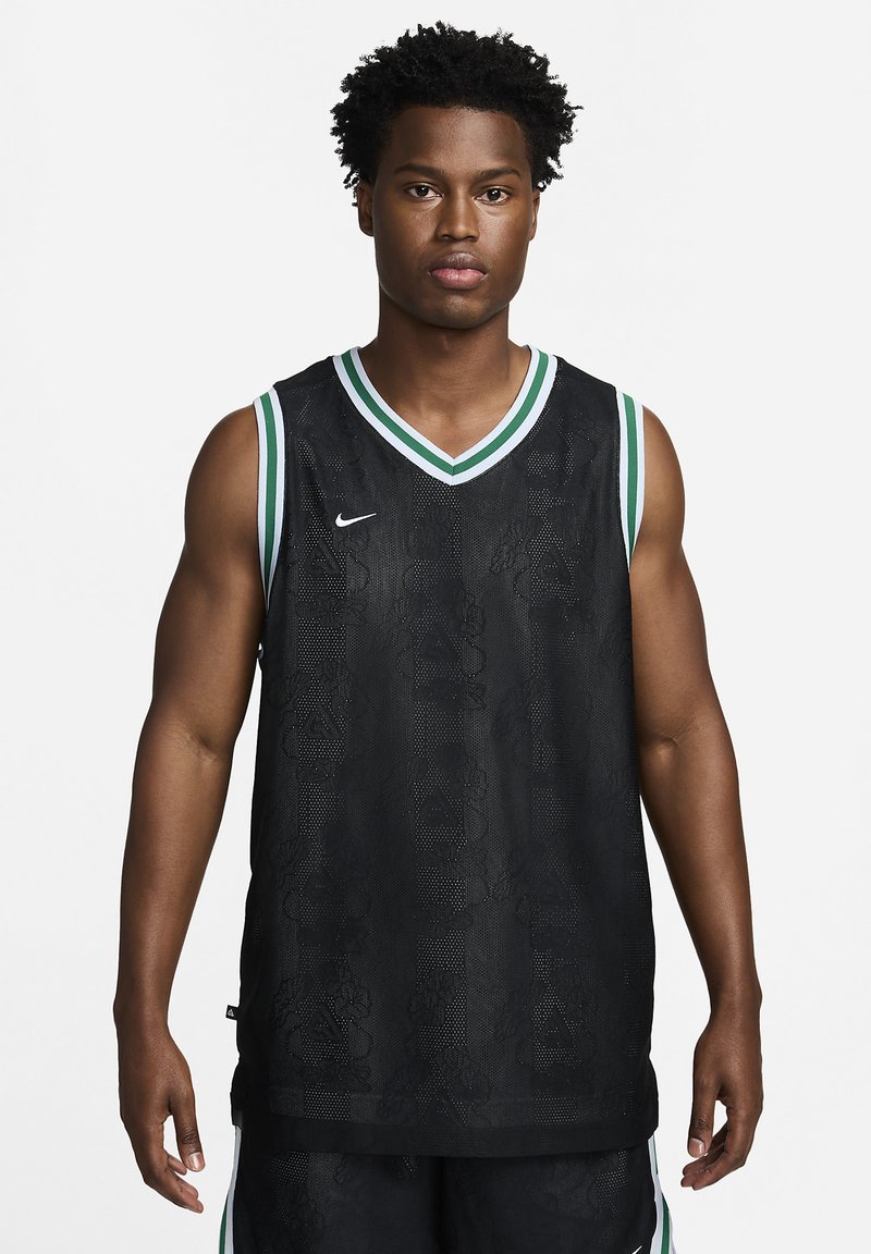 Hombre de pie mirando hacia adelante, vestido con una camiseta de baloncesto negra con adornos verdes y blancos, y pantalones cortos negros a juego.
