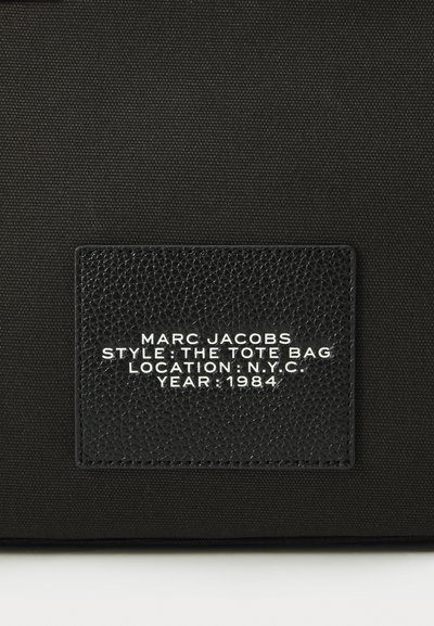 Black tote bag featuring a textured leather label with white embossed text: "MARC JACOBS STYLE: THE TOTE BAG LOCATION: N.Y.C. YEAR: 1984."