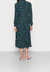 Kvinna i en långärmad, mörk blommig midi-klänning med flerfärgade blommor, stående barfota med vita sandaler på ett ljust golv.