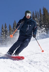 De skispullen omvatten rode ski's, zwarte skiërschoenen, een blauwe jas, donkere broek, oranje skistokken en een groene helm op de skiër. Sneeuwachtige achtergrond.