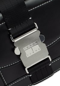Bolso de imitación de cuero negro con herrajes plateados, que presenta una hebilla texturizada y acentos de costura. Incluye una correa de webbing negra resistente.