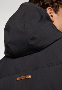 Sort hættejakke lavet af tekstureret stof. Har et læderlogo-patch og broderede detaljer på skuldrene.