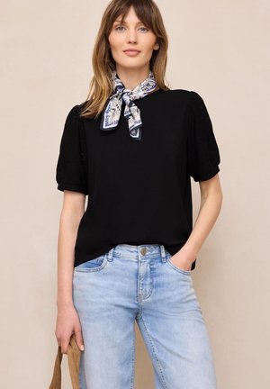 Kobieta o jasnobrązowych włosach ubrana w czarną bluzkę z bufkami, niebieską, wzorzystą chustę zawiązaną na szyi, jasnoniebieskie dżinsy, trzymająca brązową torebkę.